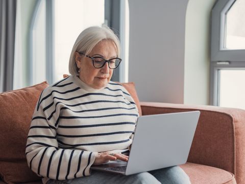 Frau mit grauen Haaren sitzt auf der Couch und nutzt den Laptop.