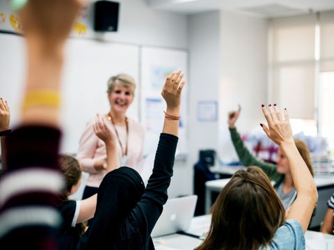 Schülerinnen und Schüler sitzen in einem Klassenzimmer und melden sich. Die Lehrerin steht vor dem Tafel.