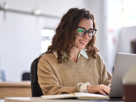 Junge Frau sitzt lächelnd vor dem Laptop an einem Schreibtisch.